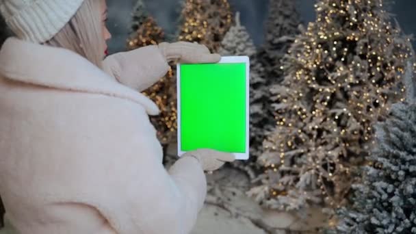 在白雪覆盖的森林里 女人手持绿色屏风装置 树上点缀着背景灯 寒假心情录像 白色保暖衣服 手套和帽子 Chroma键屏幕 — 图库视频影像