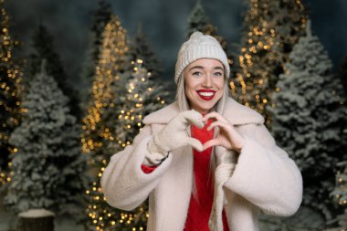 Ben de seni. Gece ormanlarında ağaçlarda ışıklar varken kış aşkı. Kış ormanındaki mutlu sarışın kadın. Kameraya bakıp gülümsüyorsun. Eller romantik sembolleri sever. Kırmızı elbiseler ve dudaklar.