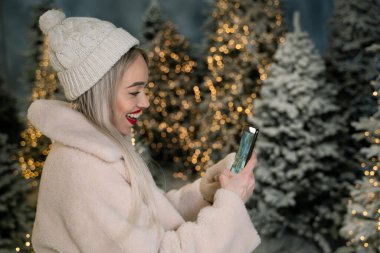 Heyecanlı mutlu kadın cep telefonuyla kış ormanı ağaçlarının fotoğrafını çekiyor. Akşam doğa yürüyüşü. Güzel sarışın kız. Kış tatillerinde blog yazarı. Sosyal medya için içerik yaratıyor.