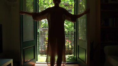 atmosphere of an optimistic future mood good morning video footage. young blonde slim woman opens the door and exits dark room onto a balcony filled with sunlight. green trees outside