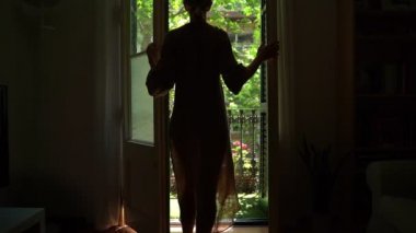 young blonde slim woman opens the door and exits dark room onto a balcony filled with sunlight. green trees outside. Welcomes the new day. Long light beige casual dress. Relaxed mood video footage