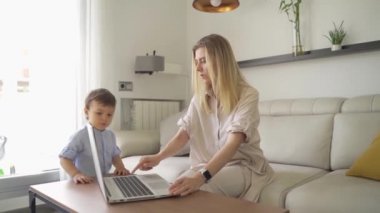 Dizüstü bilgisayarda yazan sarışın kadın çocuğa bilgisayarın nasıl kullanılacağını gösteriyor. oyun oynamak ve temel şeyleri öğrenmek. Gelecekteki programcı. Parlak oturma odası. Yavaş çekim video görüntüleri. Evde yeni yürümeye başlayan bebekle çalışan bir anne.