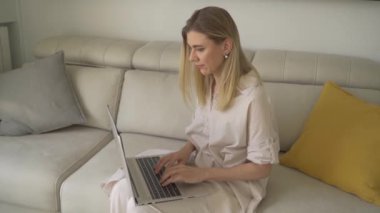 Dizüstü bilgisayar kullanan genç sarışın kadın çalışıyor, internetten alışveriş yapıyor. Yavaş çekim video görüntüleri. Kanepeli bej renkli oturma odası. Doğal gün ışığı