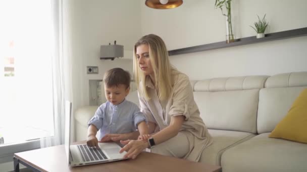 幼児の少年はラップトップで遊んでいる キーボード入力を押します ママは興奮してコンピューターを持って赤ん坊を見ている 将来のプログラマー 家で働くブロンドの女性は 日当たりの良い部屋に座ってコンピュータを使用して — ストック動画
