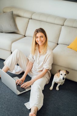 Laptop kullanan güzel sarışın kadın. Evden çalışıyor, internetten alışveriş yapıyor. Freelancer ya da uzaktan kumanda. Oturma odasında küçük sevimli bir köpekle otururken. Dikey bileşim