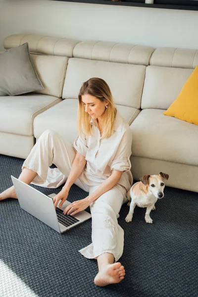 在家工作 快乐的金发碧眼的女人用笔记本电脑打字 在阳光灿烂舒适的客厅里 坐在地板上 靠着米黄色的沙发 远程办公工作 — 图库照片