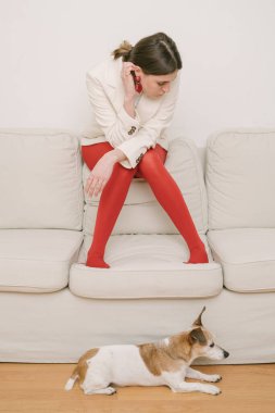 Kırmızı taytlı güzel bir genç kız bej bir kanepede başı eğik oturuyor. Düşünceli üzgün poz. Aşağı bakıyor. Küçük köpek Jack Russell terrier yerde oturuyor. Kompozisyon kavramı. Stres havası