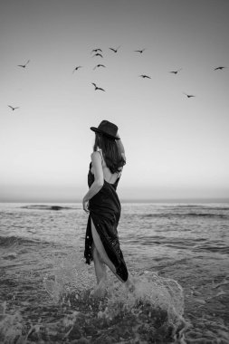 Ayrılma konsepti. Uzun siyah elbisesi ve şapkası olan romantik, ince bir kız gidiyor. Arkasında dalgalar olan deniz. Romantik dikey siyah beyaz kompozisyon. Geçmişe özlem