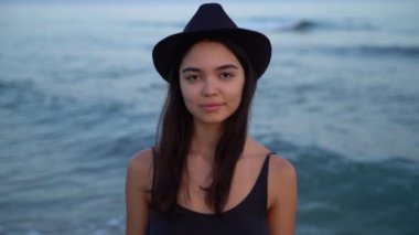 Siyah şapkalı esmer kız. Arka planda deniz resmi olan yakın plan. Duygular ve anılar konsepti. Dişi düşünceli ince kız kameraya bakıyor. Mavi günbatımı yumuşak ışık video görüntüleri