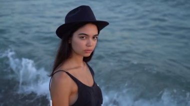 young brunette beautiful woman in hat in profile in front of sea. Romantic Slow motion video footage. disturbing anxious haughty eyes look movement of the waves on the background. 