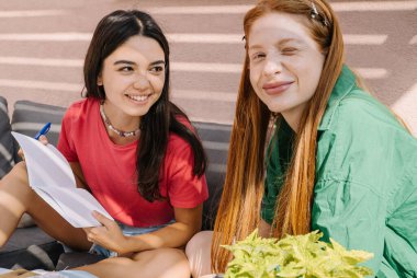 Two girls looking at camera and smiling. Studying together, making notes, preparing for exam. Back to school theme. summer sunny terrace. Green and red colors. Brainstorm team work. joint discussion