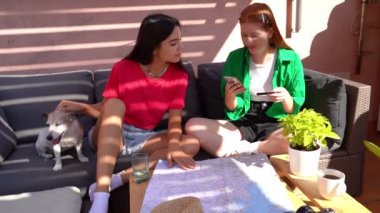 two girls friends at summer sunny terrace with striped sunlight shade booking tickets online paying with card using cell phone. Online payment theme video footage. Planning travel table with map 