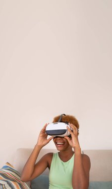 Exited black skin young woman wearing virtual reality simulator siting on sofa room at home. Happy smiling positive emotions. touches glasses with hands. Long vertical banner composition. copy space