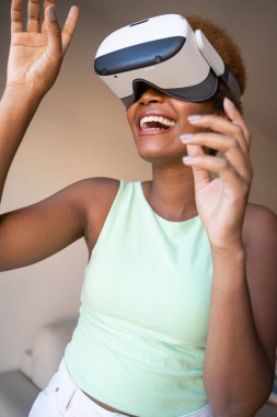 Black skin young woman wearing virtual reality Headset simulator  playing VR game. green colors clothes. beige background. smiling exited surprised emotion. Vertical composition