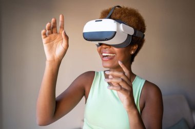 Black skin young woman wearing virtual reality simulator gesturing while siting on sofa in living room at home