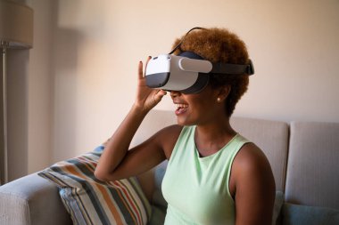 VR Headset wearing young black woman using at home sitting on sofa. smiling surprised emotion. happy positive experience. Horizontal composition. 