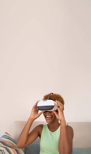 Exited black skin young woman wearing virtual reality simulator siting on sofa room at home. Happy smiling positive emotions. touches glasses with hands. Long vertical banner composition. copy space