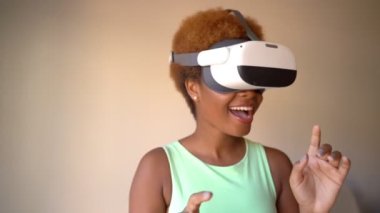 Excited surprised Black woman testing wearing virtual reality simulator headset home. Happy positive emotions. green clothes. touches glasses with hands. Video footage green clothes beige background