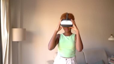 Black woman wearing virtual reality simulator siting on sofa in  at home. Happy positive emotions. green clothes. touches glasses with hands. video footage. green clothes beige background