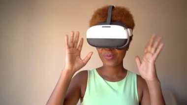 Excited surprised Black woman testing wearing virtual reality simulator headset home. Happy positive emotions. green clothes. touches glasses with hands. Video footage green clothes beige background
