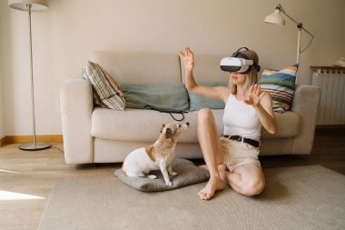 Sevimli köpek şaşkınlık içinde sanal gerçeklik simülatörü kullanan genç, narin sarışın kadının elleri havada yerde oturmasını izliyor. VR gözlükleri 360 derecelik video izliyor.