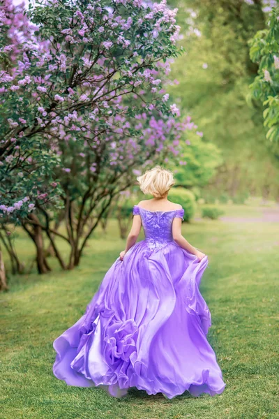 girl in a long lilac, blue dress runs in the park. Photo from the back. Lilac flowers grow in the park