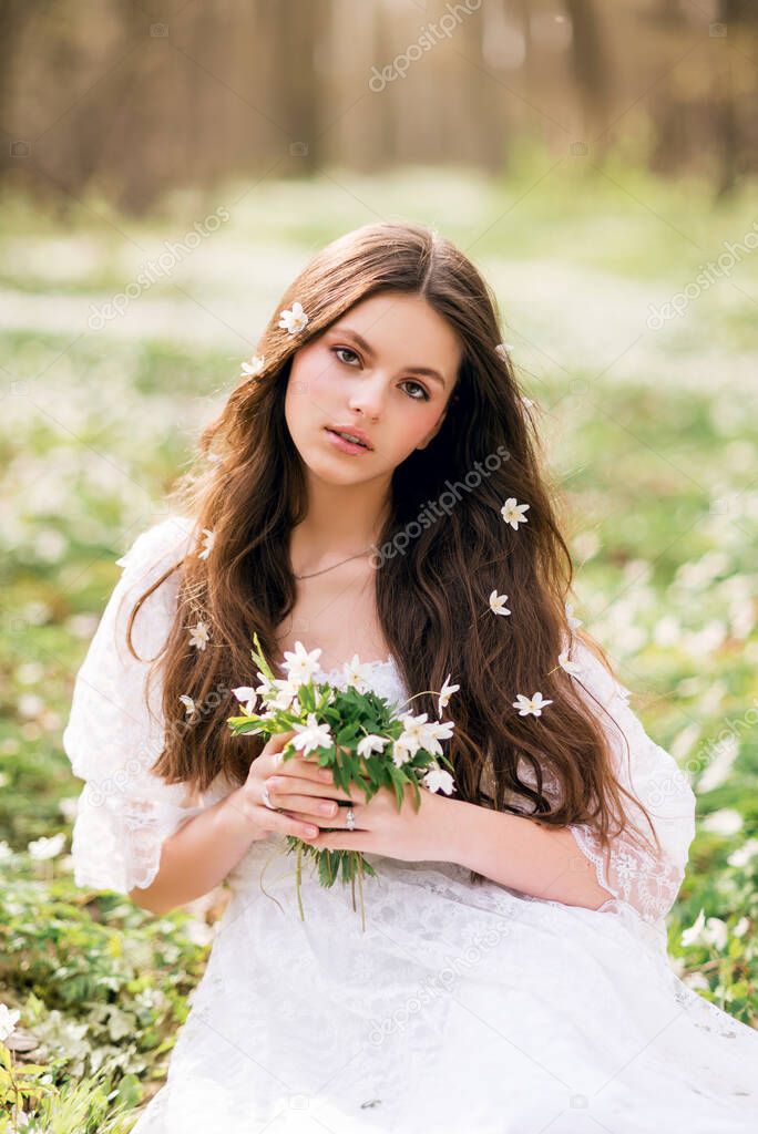 Portrait of a young woman in a white dress with primroses. A girl in the spring forest. White anemones in long brown hair..