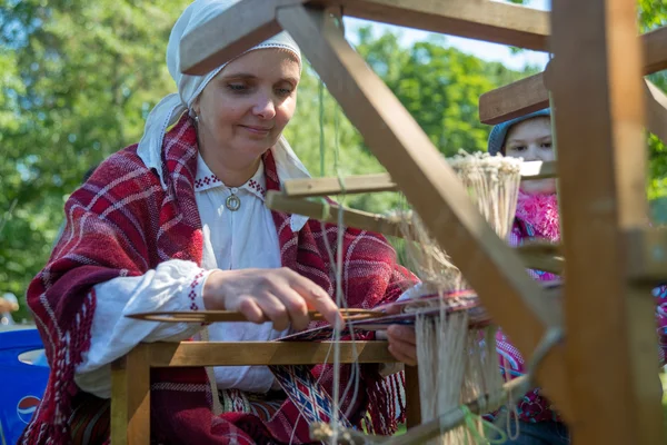 Mujer mayor sonriente que trabaja en el telar de tejer estudia a los niños. Ropa étnica tradicional del Báltico . — Foto de Stock