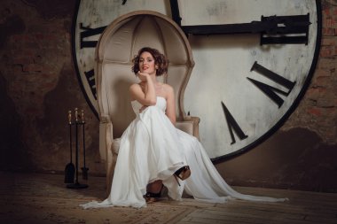 A smiling girl in a wedding dress in strange chair. The bride in a chair on the background of clocks and fireplace tool set. Horizontal clipart