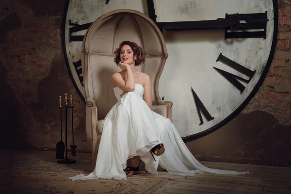 一个微笑的女孩穿婚纱的样子在奇怪的椅子上。新娘在坐在椅子上的时钟和壁炉工具的背景设置。水平 — 图库照片