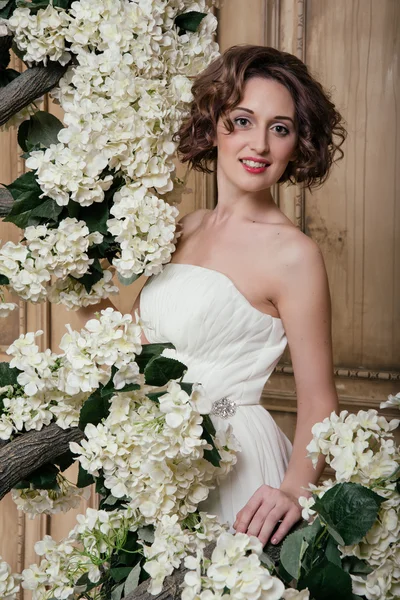 Noiva sorridente rodeada de flores brancas. Retrato de morena sorridente em vestido branco, cercada por flores brancas . — Fotografia de Stock