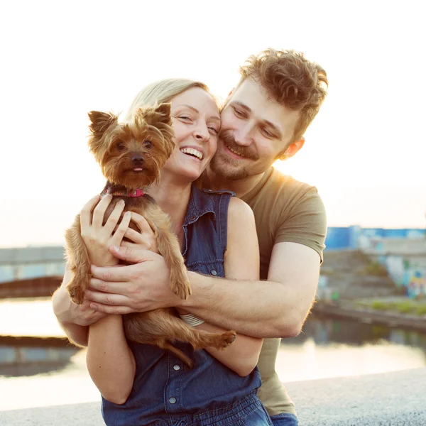 Riendo pareja joven con perro pequeño en la luz del atardecer — Foto de Stock