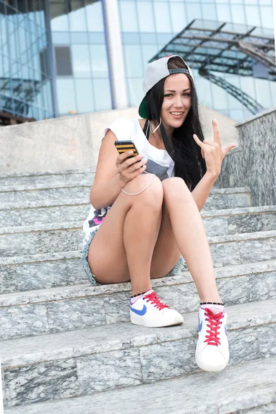 Jovem adolescente com fones de ouvido, cabelo longo preto depois da escola sentado em passos de shopping center com seu smartphone — Fotografia de Stock