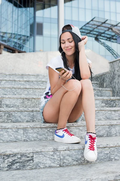 Jovem adolescente com fones de ouvido, cabelos longos pretos e jeans sentados em degraus, sorrindo e ouvindo a música em seu smartphone . — Fotografia de Stock