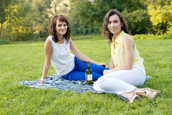 Two young pretty girlfriends open a bottle of wine on the meadow during the picnic and sitting on the blanket. — Stock Photo, Image