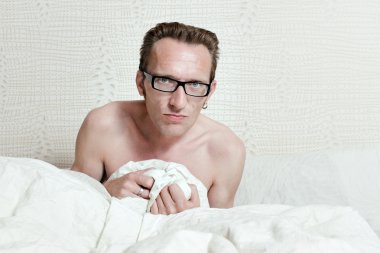 Irritated skinny naked man with angry face sitting in the bed in white blanket and looking into the camera. clipart