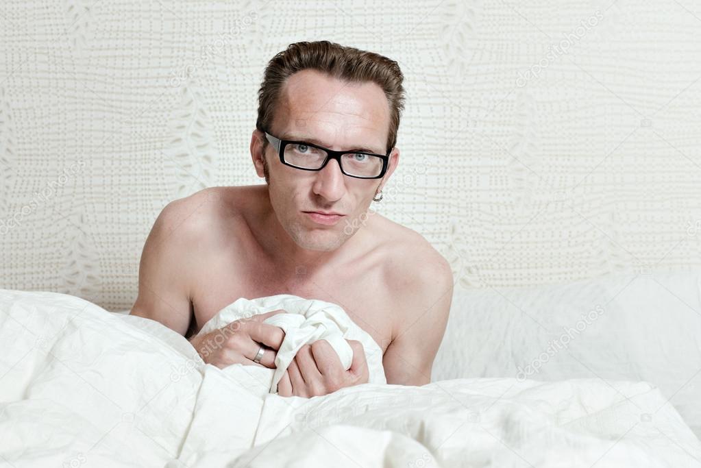 Irritated skinny naked man with angry face sitting in the bed in white blanket and looking into the camera.