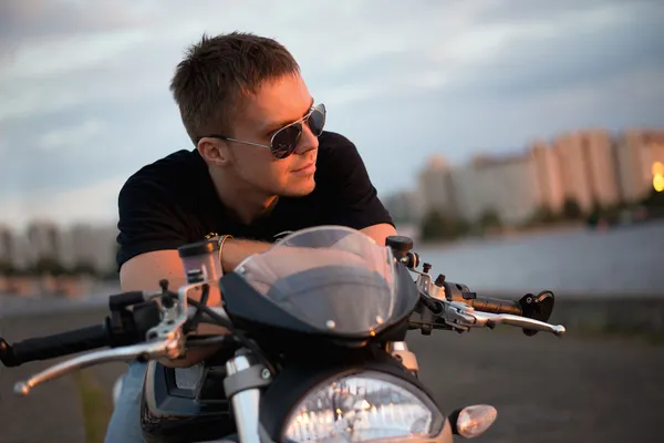 Retrato romântico homem motociclista bonito em óculos de sol senta-se em uma bicicleta em um pôr do sol perto do lago e da cidade — Fotografia de Stock