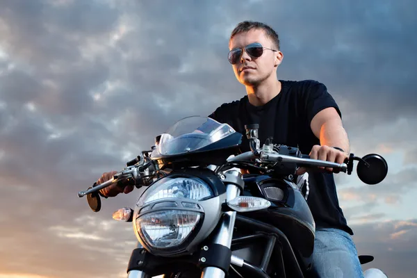 Retrato romântico homem motociclista bonito em óculos de sol senta-se em uma bicicleta em um pôr do sol perto do lago — Fotografia de Stock