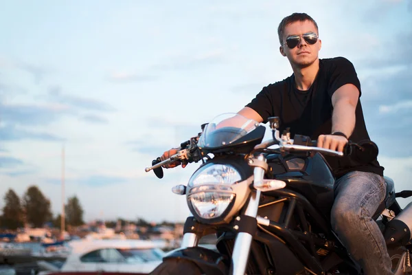 Ritratto romantico bell'uomo motociclista in occhiali da sole si siede su una bicicletta su un tramonto vicino al lago — Foto Stock