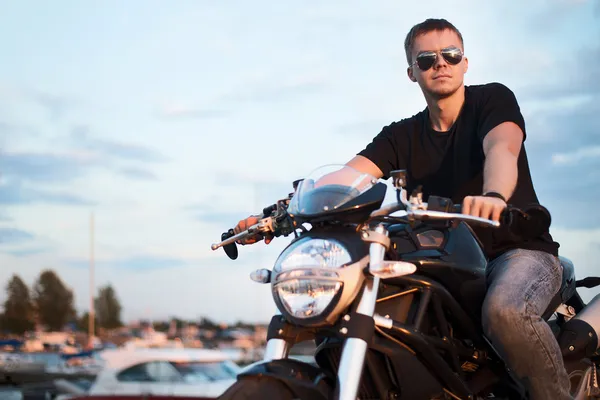 Romántico retrato guapo motorista en gafas de sol se sienta en una bicicleta en una puesta de sol cerca del lago — Foto de Stock