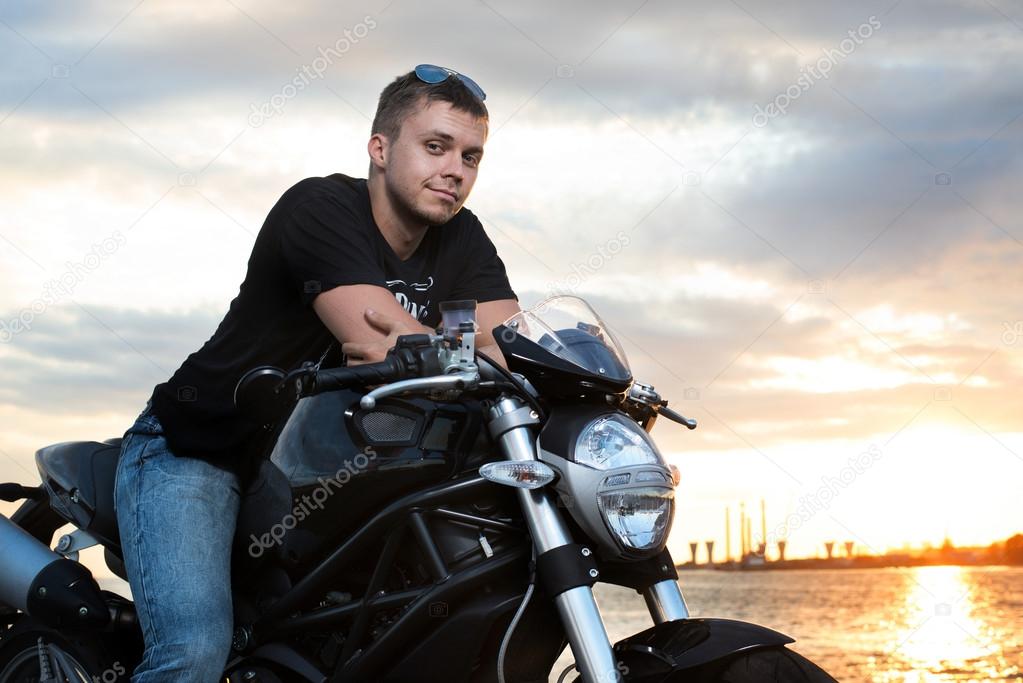 Romantic portrait handsome biker man in sunglasses sits on a bike on a sunset near lake