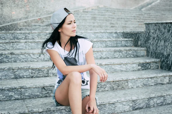 Young teenage girl with headphones,black long hair and jeans shorts sitting on steps and attentively looking right. — Stock Photo, Image