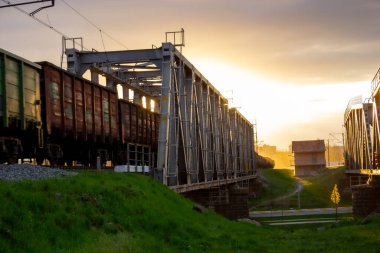 A railway bridge on which a train with wagons moves against the background of a bright sun.