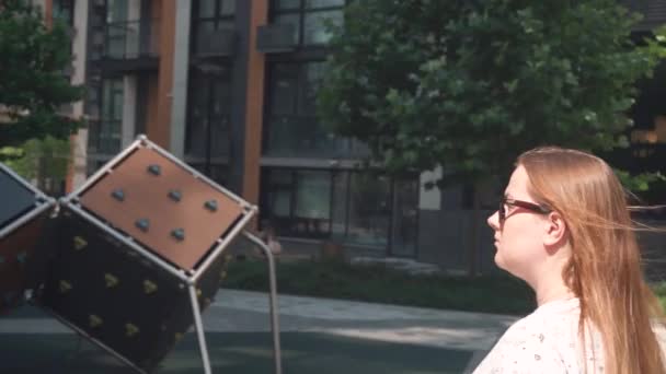 Een meisje met bruin haar in een zonnebril loopt door een woonwijk, draait haar hoofd naar de camera — Stockvideo