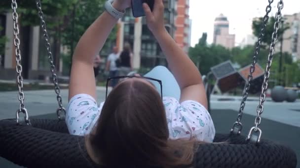 Una joven casada con gafas de sol se encuentra en un columpio casero y escribe un mensaje en el teléfono. — Vídeo de stock