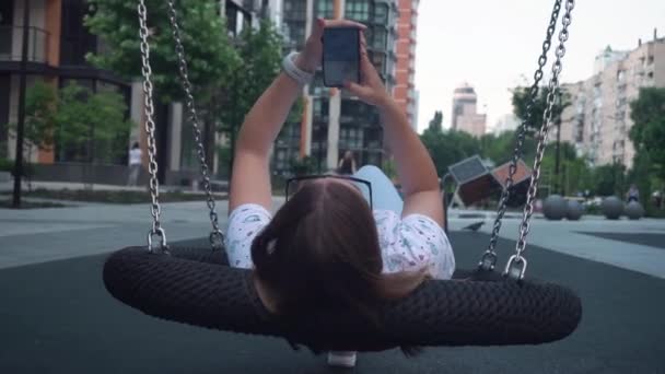 Ένα νεαρό παντρεμένο κορίτσι με γυαλιά ηλίου ξαπλώνει σε μια σπιτική κούνια και γράφει ένα μήνυμα στο τηλέφωνο. — Αρχείο Βίντεο