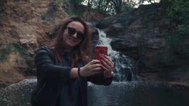 Şirin, şık bir kız, gülümsüyor, güneş gözlükleriyle poz veriyor, deri ceket, saat, yüzük, selfie çekiyor, blog için video çekiyor, arka plandaki küçük şelaleye karşı. Ağır çekim. Bahar, yaz günü doğa taşları