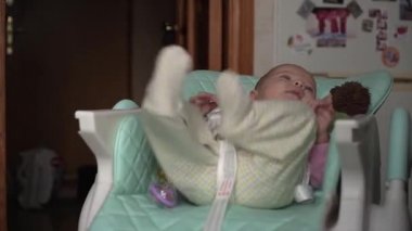 Küçük, tatlı bir bebek, mutfakta çocuk koltuğunda sıkılıyor. Titriyor, hareket ediyor, bacaklarını, kollarını hareket ettiriyor, etrafa bakıyor, sıkıntıdan titriyor. Kamera sola doğru sorunsuz hareket ediyor.