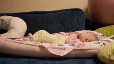 Neşeli küçük bir bebek, ellerini ve parmaklarını ağzına koyar, tadına bakar, tek başına oynar, eğlenir, odayı inceler. Çocuk tulumu giymiş bir çocuk, koza içinde bir yatağın üzerinde. Yan görünüm